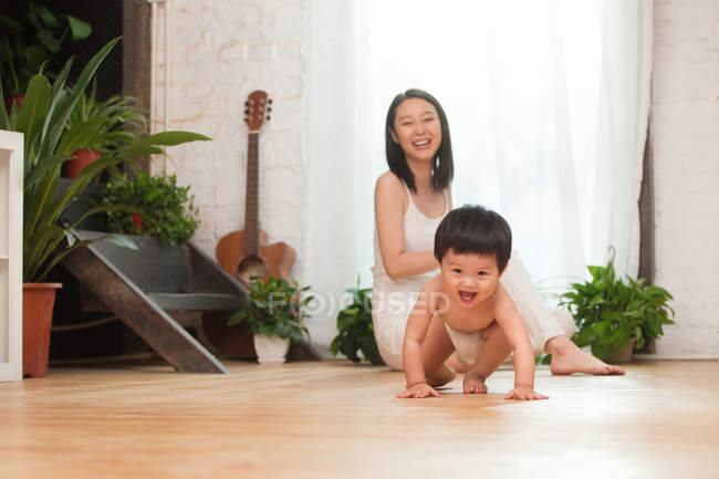 Feliz joven madre mirando a su adorable bebé excitado arrastrándose en el suelo en casa - foto de stock