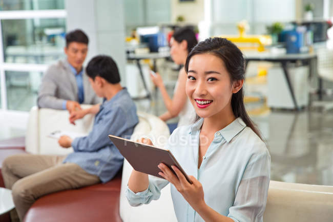 Красивая молодая азиатская бизнесвумен, использующая цифровой планшет и улыбающаяся в камеру, в то время как коллеги работают в офисе — стоковое фото