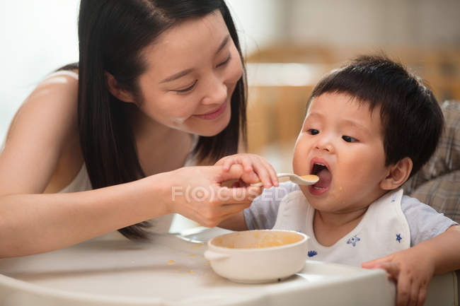 Sorridente giovane madre che tiene cucchiaio e nutre il suo adorabile bambino a casa — Foto stock