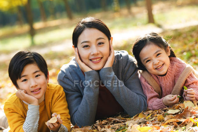 Lächelnde asiatische Mutter mit Tochter und Sohn auf Laub im herbstlichen Park liegend und in die Kamera blickend — Stockfoto