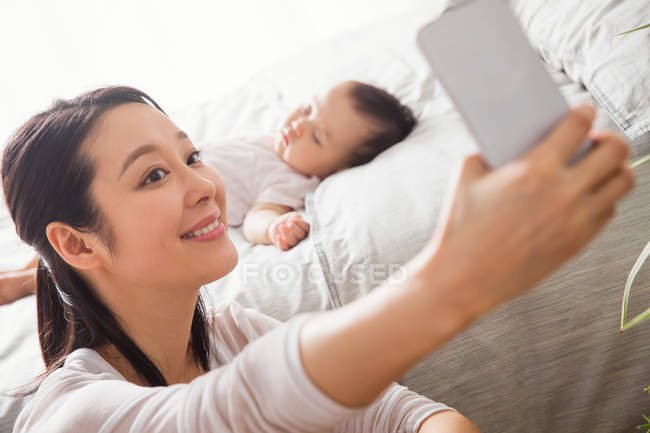 Selective focus of young mother taking selfie with smartphone while baby sleeping on bed — Stock Photo