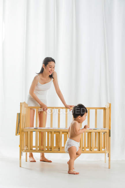 Smiling young mother looking at cute toddler in diaper standing near crib at home — Stock Photo