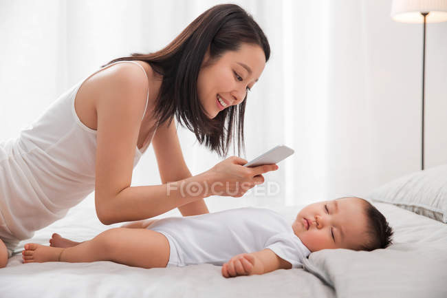Улыбающаяся молодая азиатская женщина с смартфоном и фотографирующая очаровательного ребенка спящего на кровати — стоковое фото