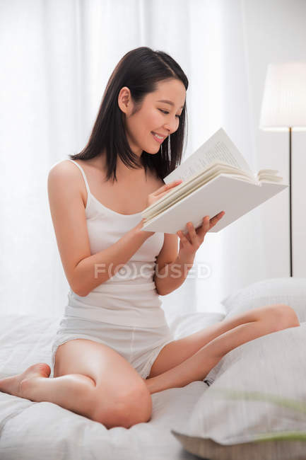 Красивая улыбающаяся молодая азиатская женщина сидит на кровати и читает книгу — стоковое фото