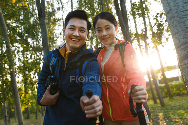 Feliz joven asiático pareja con binoculares y trekking palos sonriendo a cámara en bosque - foto de stock