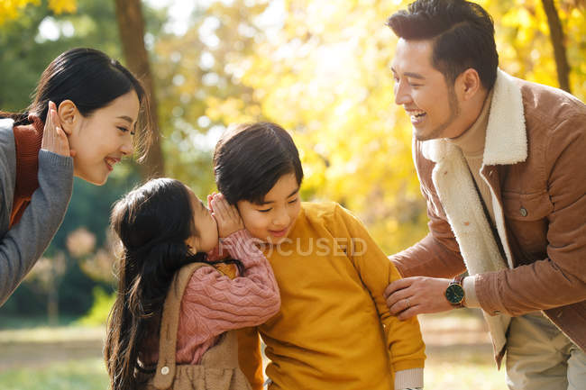 Sonrientes padres jóvenes mirando a los hermanos susurrando algo en el parque de otoño - foto de stock