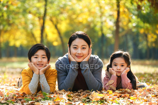 Sorridente asiatica madre con figlia e figlio sdraiato sul fogliame nel parco autunnale e guardando la fotocamera — Foto stock