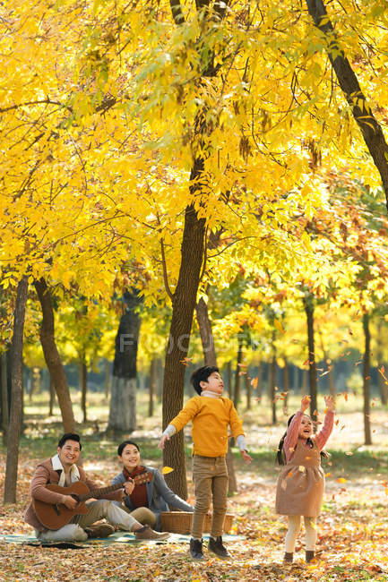 Adorable happy kids playing with autumn leaves and parents enjoying guitar during picnic in forest — Stock Photo