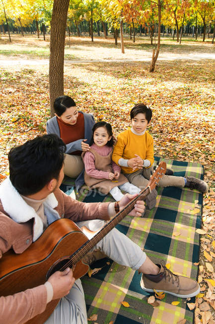 Високий кут зору батька, який грає на гітарі та щасливій сім'ї, що сидить на площині в осінньому парку — стокове фото