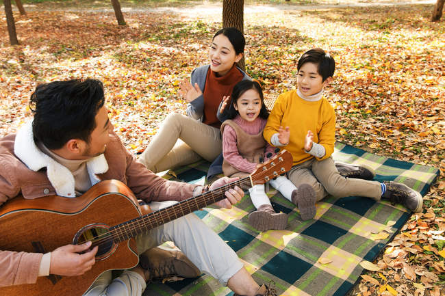 High angle view of smiling father playing guitar and happy family sitting on checkered plaid in park — Stock Photo