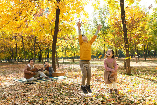 Niños felices jugando con hojas de otoño mientras los padres descansan en cuadros a cuadros en el parque - foto de stock