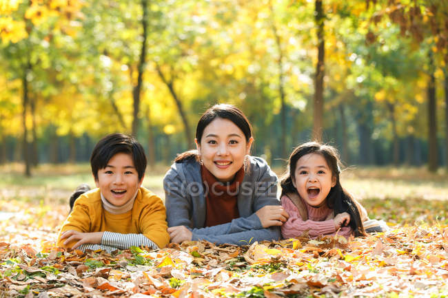 Sonriente asiático madre con hija e hijo acostado en follaje en otoñal parque y mirando a cámara - foto de stock