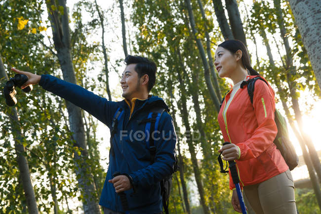 Low angle view of happy young asian couple with trekking sticks looking away in forest — Stock Photo