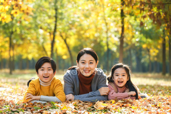 Smiling asian mother with daughter and son lying on foliage in autumnal park and looking at camera — Stock Photo