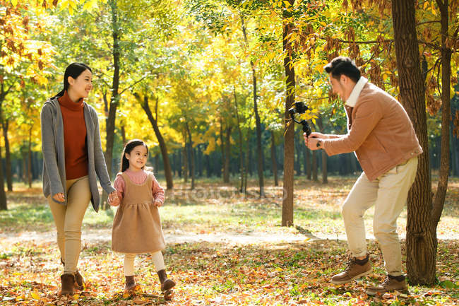 Счастливый азиатский отец фотографирует дочь и жену со смартфоном в осеннем парке — стоковое фото