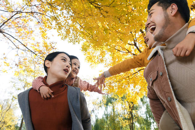 Low angle view of happy young parents piggybacking adorable kids in autumn forest — Stock Photo