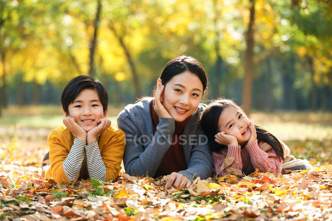 Улыбающаяся азиатская мать с дочерью и сыном, лежащими на листве в осеннем парке и смотрящими в камеру — стоковое фото