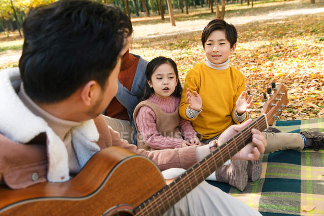 Mutter mit entzückenden Kindern schaut Vater beim Gitarrespielen im Wald an — Stockfoto