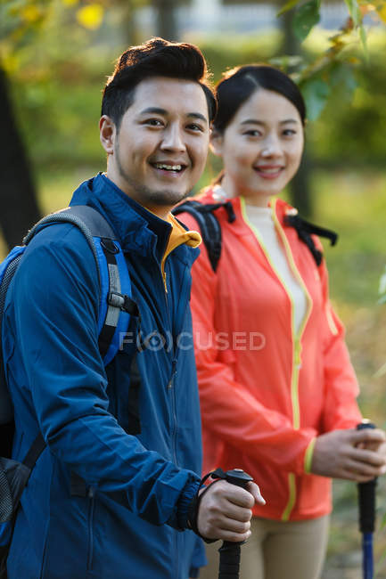 Feliz joven asiático pareja con mochilas y trekking palos sonriendo a cámara en bosque - foto de stock
