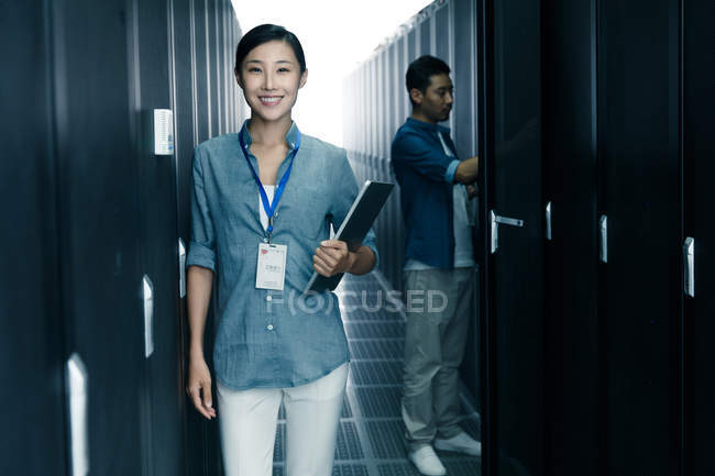 Женщина-работница улыбается в камеру, в то время как мужчина-инженер работает в комнате технического обслуживания — стоковое фото