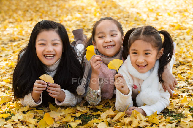 Three adorable smiling asian kids lying on yellow foliage and holding leaves in autumnal park — Stock Photo