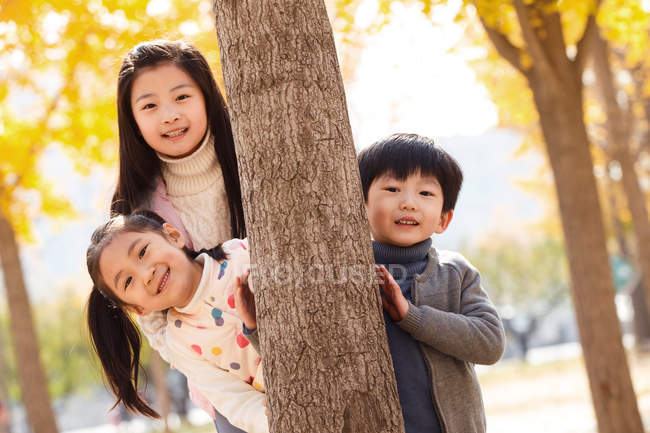 Happy boy and girls standing together near tree and smiling at camera in autumn park — Stock Photo