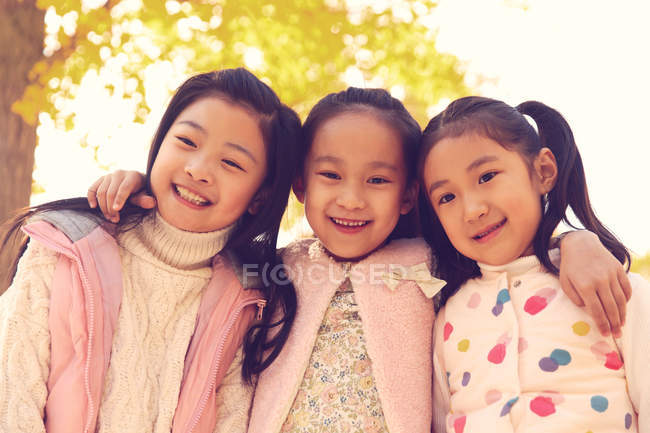 Niedrigwinkel-Ansicht von drei entzückenden lächelnden asiatischen Kindern, die sich im herbstlichen Park umarmen und in die Kamera schauen — Stockfoto