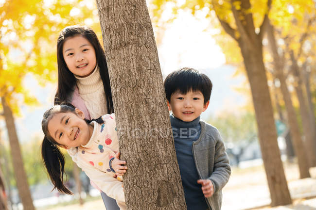 Щасливий хлопчик і дівчата стоять разом біля дерева і посміхаються на камеру в осінньому парку — стокове фото