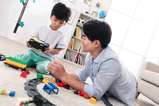 Smiling asian father and son playing with toy railway station on floor at home — Stock Photo