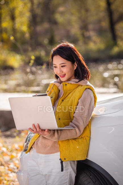 Joyeuse asiatique femme tenant ordinateur portable et penché sur la voiture en forêt automnale — Photo de stock