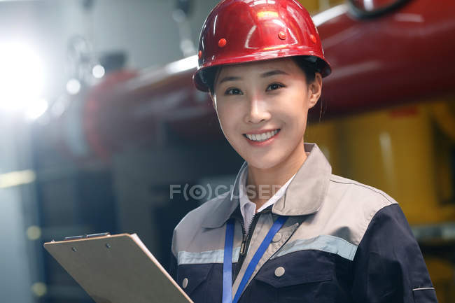 Sonriente técnico en casco inspeccionando el control de incendios en fábrica y mirando a la cámara - foto de stock
