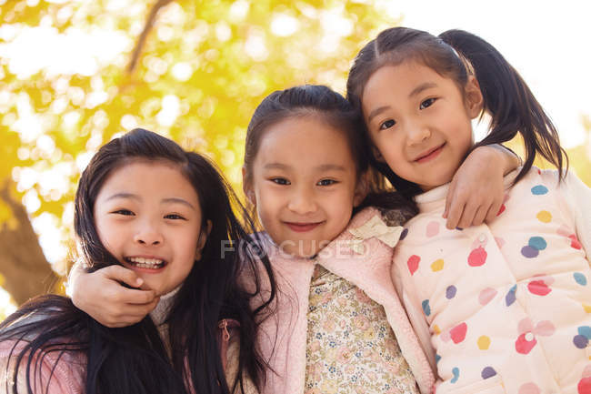 Низький кут зору трьох чарівні посміхаючись азіатських дітей обійматися в осіннього парку і дивлячись на камеру — стокове фото