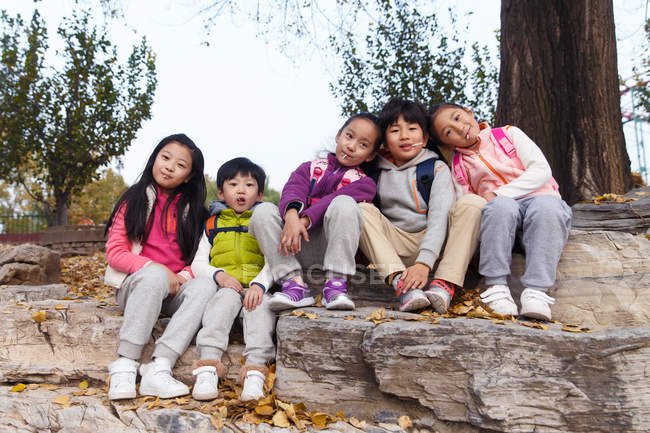 Five adorable asian kids sitting on stones and looking at camera in autumnal park — Stock Photo
