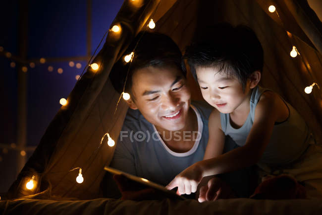Heureux asiatique père et préscolaire fils regarder quelque chose sur tablette dans tente à la maison — Photo de stock