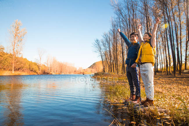Feliz casal asiático de pé com as mãos para cima perto do lago na floresta outonal — Fotografia de Stock