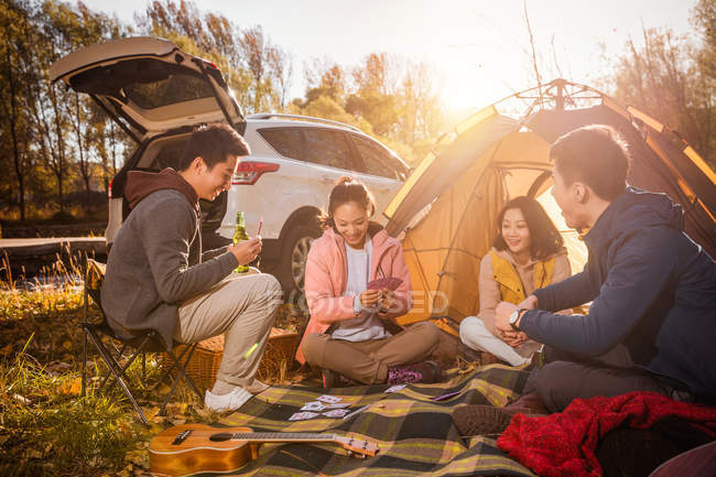 Four asian friends playing cards on blanket near tent in autumnal forest — Stock Photo