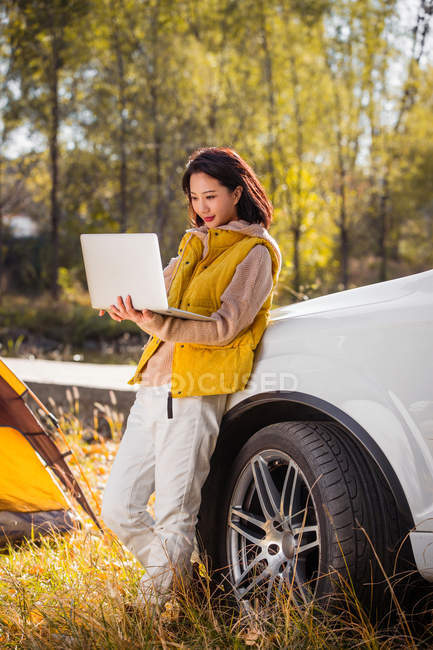 Allegro asiatico donna holding laptop e pendente su auto in autunnale foresta — Foto stock