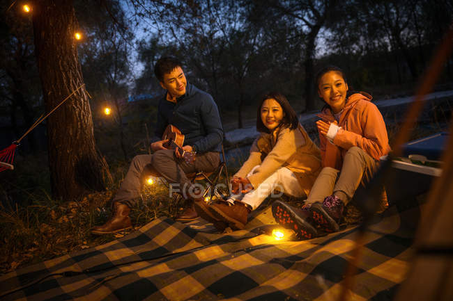 Три азиатских друга сидят на одеяле с укулеле и смотрят в камеру в осеннем вечернем лесу — стоковое фото