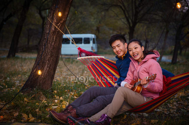 Asian girlfriend playing ukulele for boyfriend on hammock in autumnal forest — Stock Photo