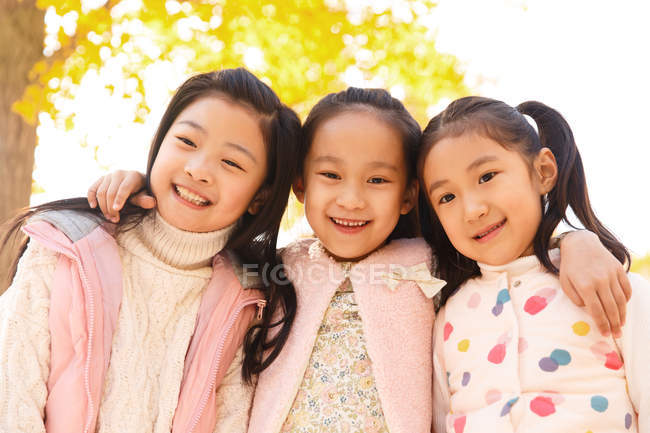 Baixo ângulo vista de três adorável sorrindo asiático crianças abraçando no outonal parque e olhando para a câmera — Fotografia de Stock