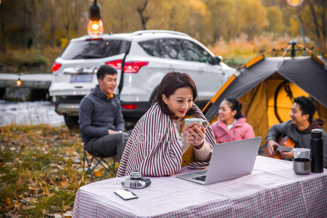 Mujer asiática sosteniendo taza de té y mirando portátil cerca de camping lugar en bosque otoñal - foto de stock