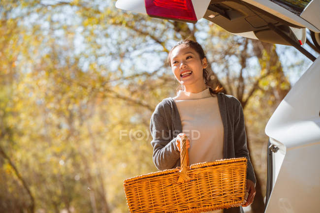 Улыбающаяся азиатка держит плетеную корзину возле машины в осеннем лесу — стоковое фото