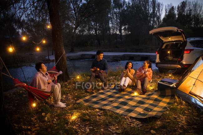 Man playing guitar for three asian friends at camping in autumnal evening forest — Stock Photo