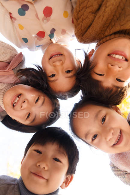 С низким углом обзора пяти очаровательных улыбающихся азиатских детей, смотрящих в камеру в осеннем парке — стоковое фото