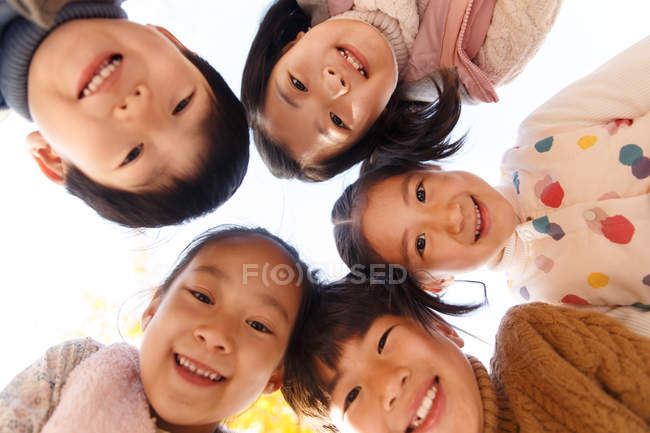 С низким углом обзора пяти очаровательных улыбающихся азиатских детей, смотрящих в камеру в осеннем парке — стоковое фото