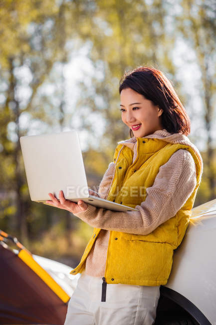 Sorridente donna asiatica che tiene il computer portatile e si appoggia sulla macchina nella foresta autunnale — Foto stock
