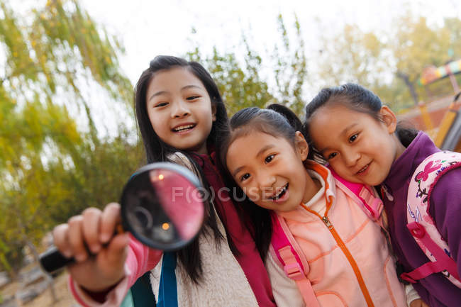 Three adorable asian kids hugging and looking at magnifying glass in autumnal park — Stock Photo