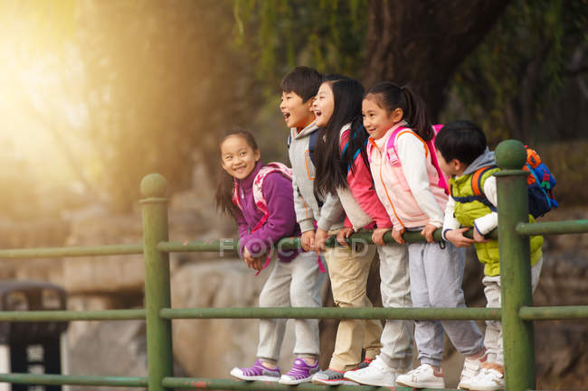 Five adorable excited asian kids leaning on fence in autumnal park — Stock Photo