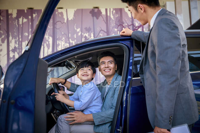 Smiling asian father and son sitting in new car and looking at car seller at showroom — Stock Photo