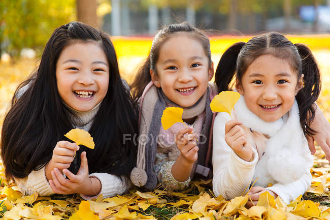 Three adorable happy asian kids lying on yellow foliage and holding leaves in autumnal park — Stock Photo